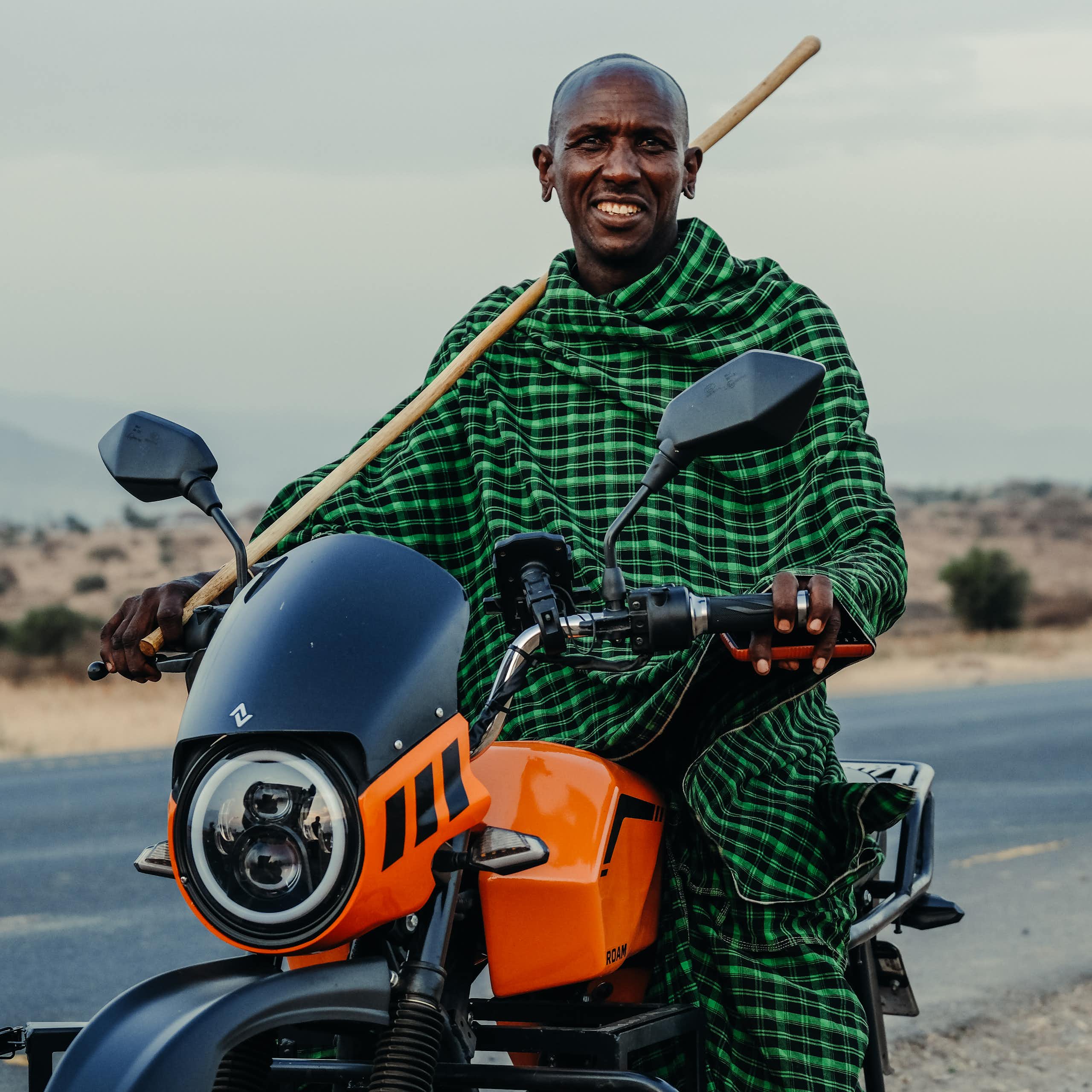 Man in checked shawl, holding a stick and sitting on a motorbike