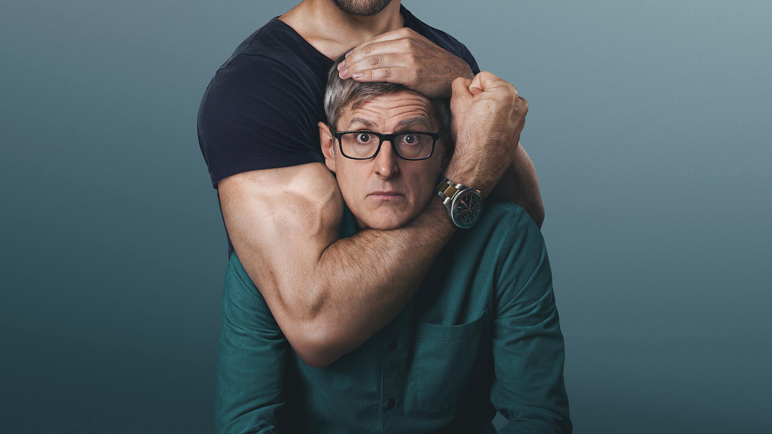 A middle-aged man in glasses stares at the camera as a muscular arm grips his head and clenches a fist.