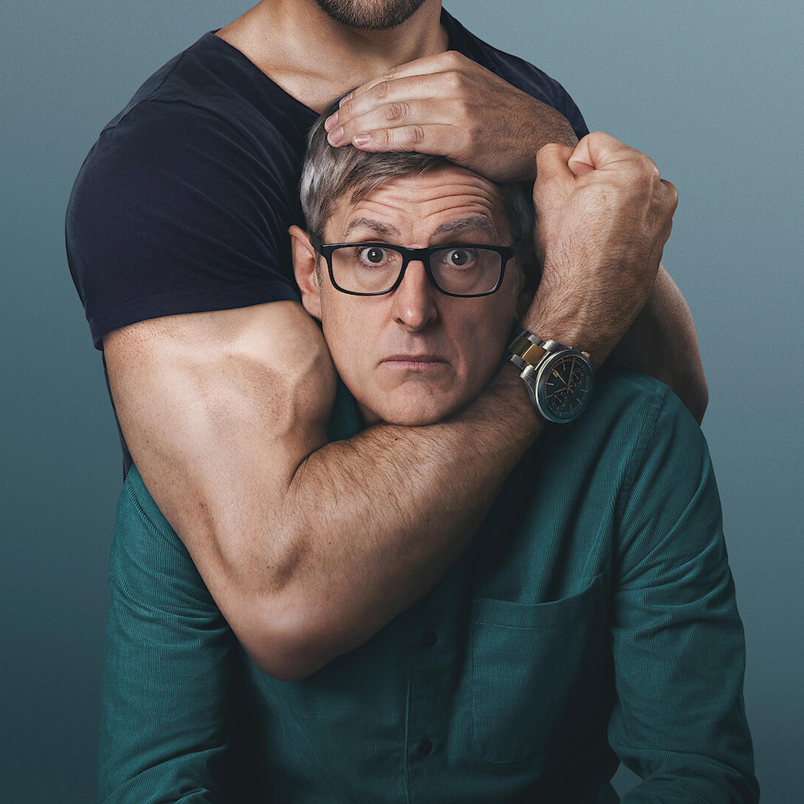 A middle-aged man in glasses stares at the camera as a muscular arm grips his head and clenches a fist.
