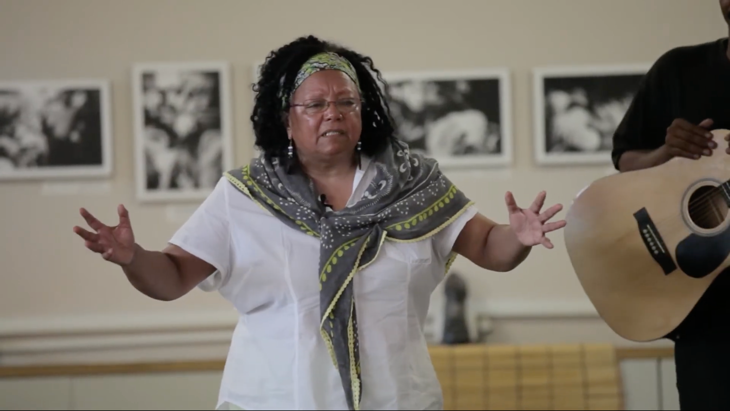 An African woman holds her arms apart in a gesture as she speaks alongside a man playing the guitar.
